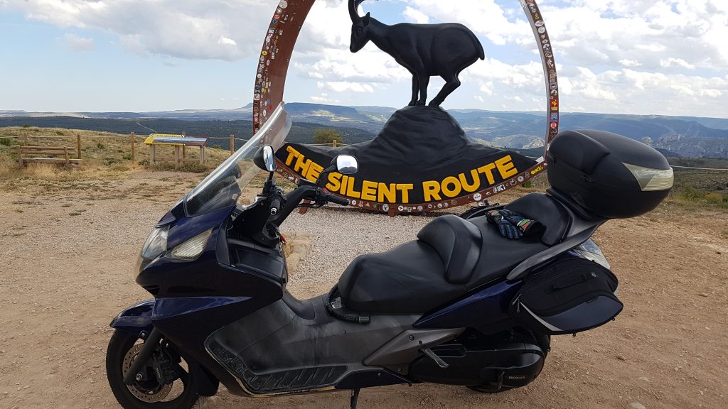 Imagen de mi moto posando delante del cartel del emblema y mascota de la ruta del silencio, y detrás de este símbolo, un paisaje precioso que me hizo echar de menos este bizcocho fácil; no el paisaje sinó el viaje.