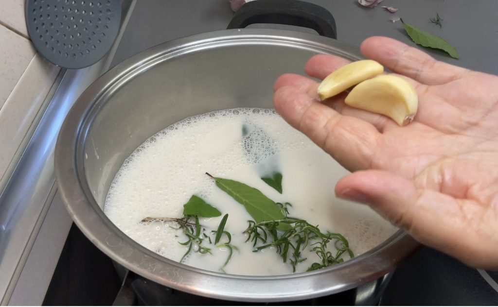 Esta imagen refleja como preparo la leche vegetal infusionada. Hay una olla al fuego con la bebida vegetal, dentro el romero y el laurel, en mi mano dos ajos pelados que me dispongo a añadirlos al líquido.