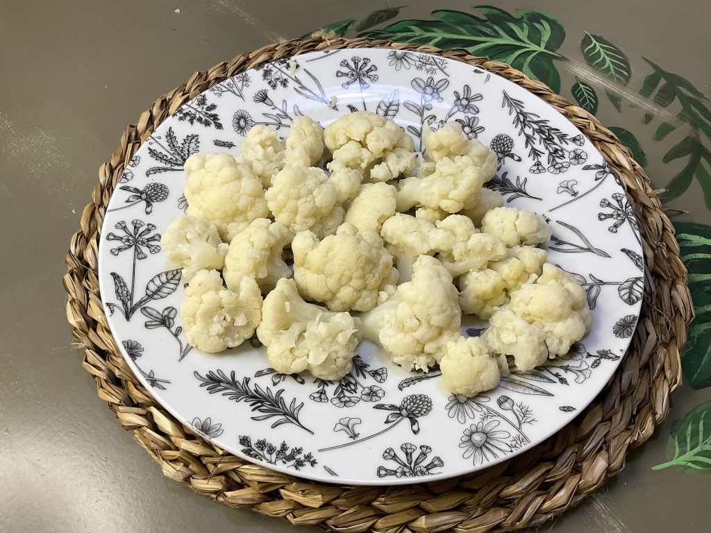 Apetitosa imagen de coliflor hervida en bebida vegetal infusionada con laurel, romero y ajos, servida en un bonito plato decorado con flores.